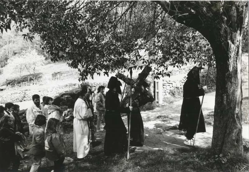 Penitentes con capirote y una cruz a la sombra de un árbol, en una fotografía en blanco y negro